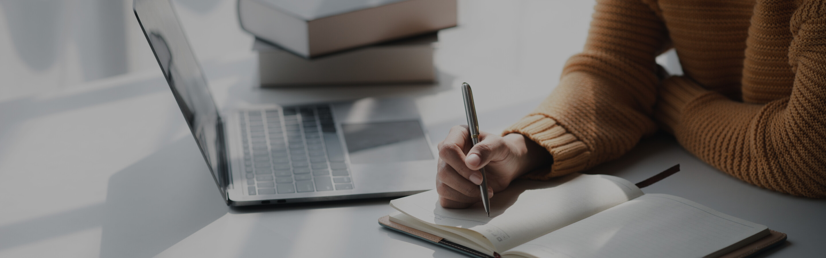 Image of a woman&#039;s arms writing in a notebook in front of a laptop