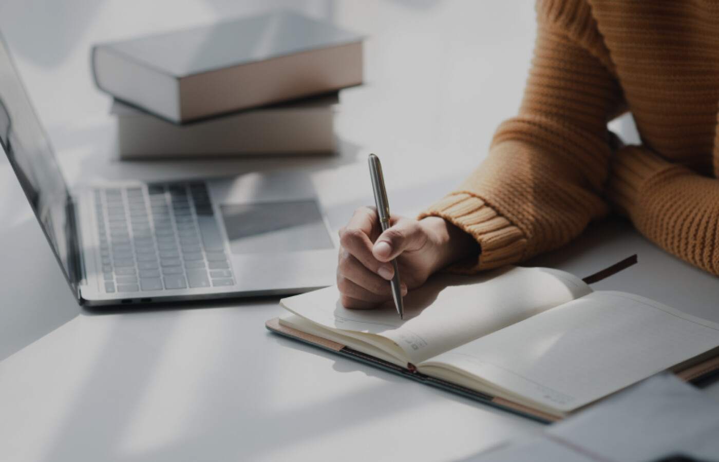 Image of a woman&#039;s arms writing in a notebook in front of a laptop