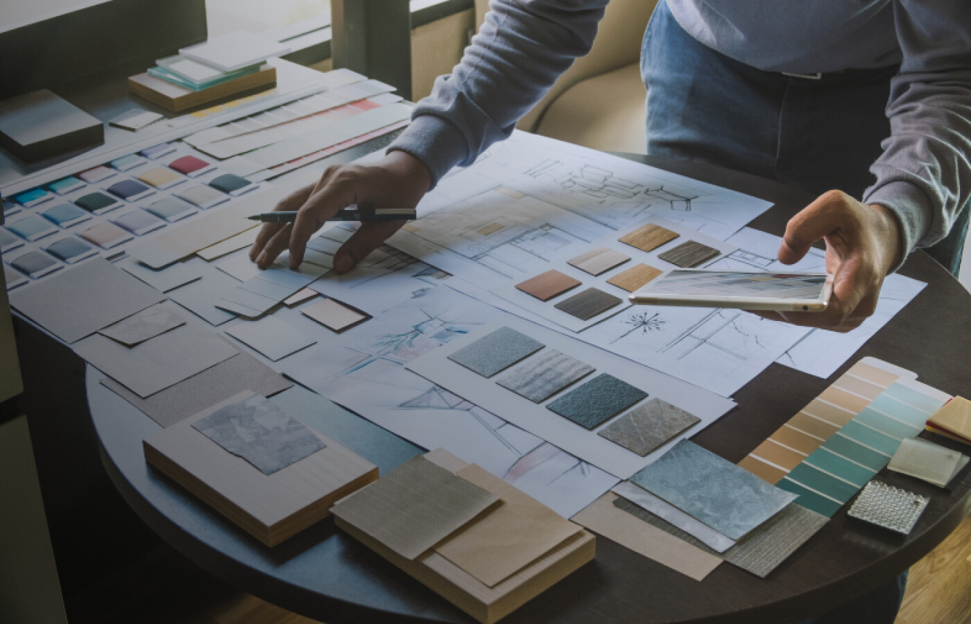 A shot of a table with interior design drawings on it and someones arm holding a pencil